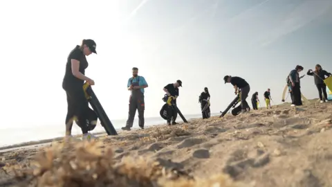 Multiple people on a beach using vacuums, viewed from ground level with sky in background.