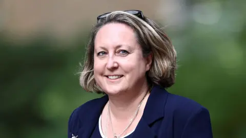 Anne-Marie Trevelyan is walking while looking beyond the camera. She has mid-length brown hair and is wearing a blue blazer.