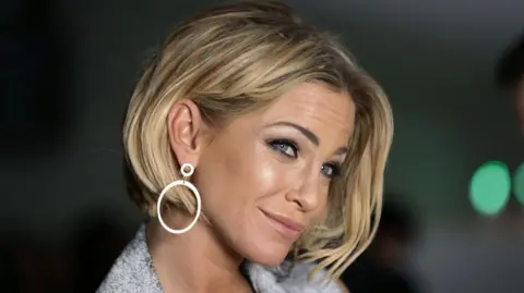 PA Media Sarah Harding, who is wearing a grey blazer and a large hooped earrings, is posing on a red carpet and she is smiling. 