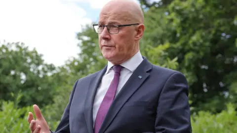 Getty Images John Swinney, who is bald with glasses, speaks outdoors with leafy green trees in the background. He is wearing a business suit. 