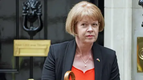 Wendy Morton stands outside the door of 10 Downing Street. She has short ginger hair and is wearing a black jacket, a red top and a necklace. 