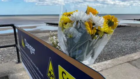 Crispin Rolfe/BBC A floral tribute close to a beach. The flowers are white and yellow and are wrapped in cellophane. The picture appears to have been taken at low tide.