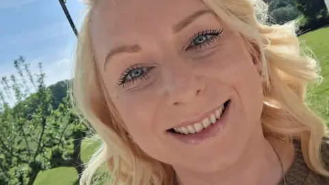 A blonde haired girl smiles at the camera. It is sunny and green in the background