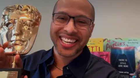 Dean Atta in a dark blue shirt, holding his Bafta award and smiling