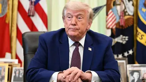 Donald Trump in the Oval Office wearing a Navy suit, white shirt and tie.