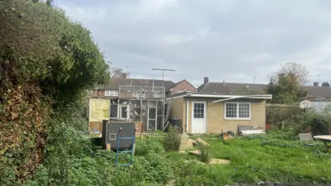 Slough Borough Council A general view of the unauthorised extension and brick outbuilding in Slough in the back garden, with appears to have overgrown grass and bushes on the left.
