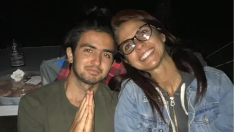 Michael Frison, left, wearing an olive coloured t-shirt. He has dark brown hair and stubble. He is holding his hands together in a prayer. He sits next to his mum Cristina, who has long dark-brown hair and black thick-rimmed glasses. She is smiling and is wearing a hoodie and a denim jacket.