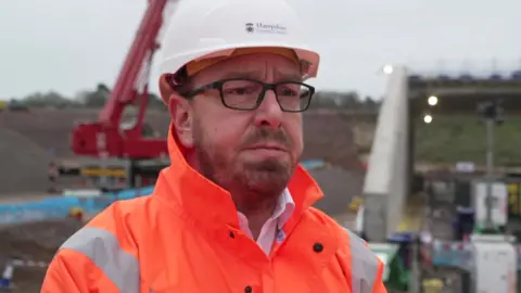 Tim Lawton looking at the camera has a white hard hat on, an orange hi-vi jacket and black framed glasses.
