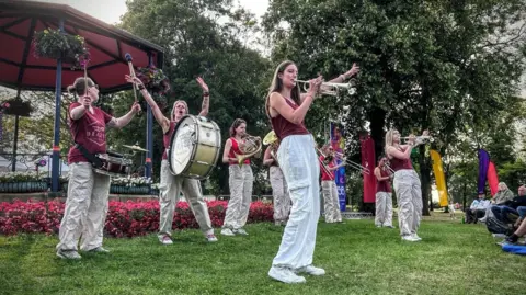 Supplied Brazen Brass Band perform in front of a park band stand at Durham Brass Festival. They all wear white trousers and red tops. 