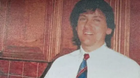 Archive photograph of Ricky Haywood smiling and wearing a white shirt and blue and red tie. He is standing in front of wood cupboards