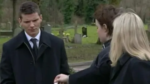 A screenshot from an old episode of EastEnders showing Sharon Mitchell handing Dennis Rickman a folded up piece of paper. They are both dressed in all black and are standing in a graveyard on a gloomy day.