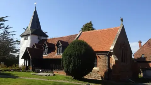 Jacqueline Eykelbosch A side-on view of St Andrew's Church, Greensted. On the left is a white-painted wooden tower and its body runs to the right, which is clad in a mix of wood, bricks and flint under a pitched roof. In front of it is grass and a yew tree.