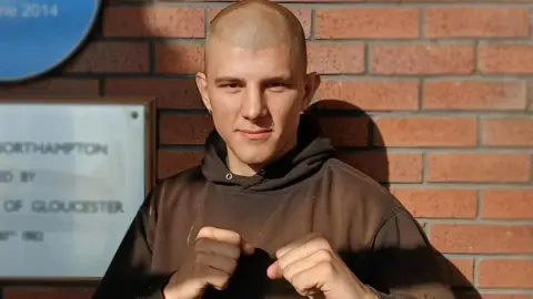 Daniel Bainger, looking at the camera, in a boxing pose. He has a nose ring, and is wearing a brown top. He is by a brick wall. He has very short shaved hair. 