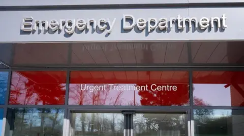 Looking up at the glass entrance to the emergency department. Above the door is a grey canopy with Emergency Department written in raised letters. Between the canopy and the top of the doors is a strip of red glass with Urgent Treatment Centre written on it in white letters. There are numerous trees behind the photographer which are reflected in the glass.
