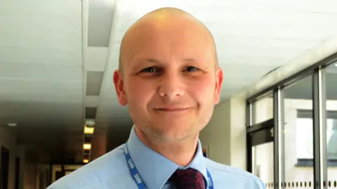 Phil Woodford who is bald with stubble wears a pale blue shirt and red tie and NHS blue and white lanyard. He is standing in a corridor close to windows and smiling.