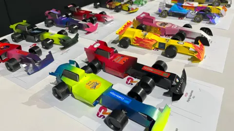 A long white rectangular table with around 20 small F1 racing cars placed on top in two rows. The cars are around the size of an A4 piece of paper and each car has at least two colours on the bodywork.