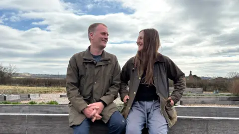 BBC James (left) and Katie Allen (right), sitting on a wooden fence smiling at each other, both wearing army green wax jackets and jeans. The farm is in the background and you can see planter-style growing areas set up behind them and lots of grass.