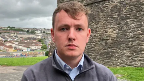 Sinn Féin assembly member Pádraig Delargy is in a grey fleece and blue shirt. He is standing beside Derry's historic walls. A number of homes can be seen in the background.
