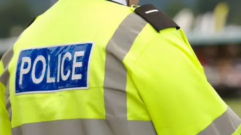 The back of a policeman who is wearing a bright yellow jacket with grey lining. There is a blue panel across the shoulders with the word "police" in white lettering