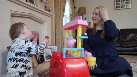 Leanne Baister and her son Toby sit in her living room playing with toys. She has long blonde hair. Toby, who has short brown hair, is offering his mother a toy ice cream. 