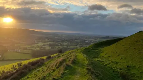 A panoramic view of the countryside with a patchwork of fields and trees stretching to the horizon. In the foreground a path can be seen winding round the side of a hill covered in green grass. The sun is setting casting a golden glow across the left-hand side of the shot.