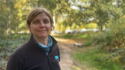 Ruth Needham stands in Beacon Hill Country Park looking at the camera. She is wearing a Trent Rivers Trust fleece and a light scarf. In the background, dips and trenches dug by the trust as part of a natural flood management scheme can be seen.