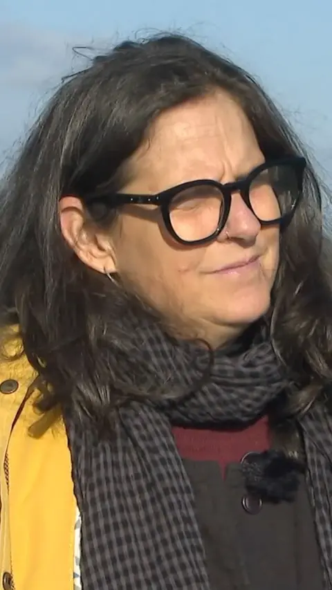 A woman with long, dark hair and black glasses wearing a yellow coat.