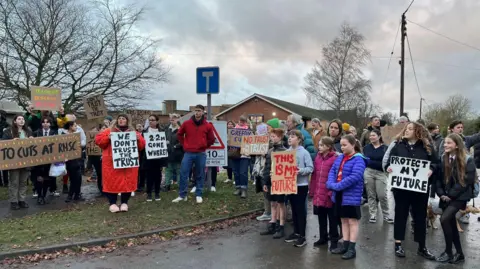 Shaun Whitmore/BBC Some 50 or so adults and children - some holding placards - stand outside a school. The placards are hand-made out of cardboard and carry messages opposing school staff cuts.