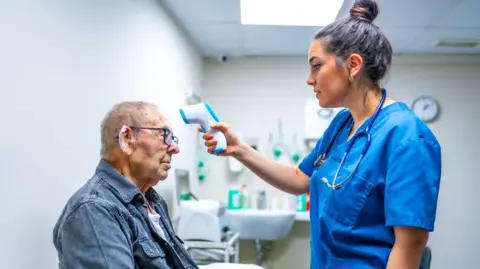 Getty Images An older man gets his temperature taken by a female doctor with blue scrubs and a hair in a bun