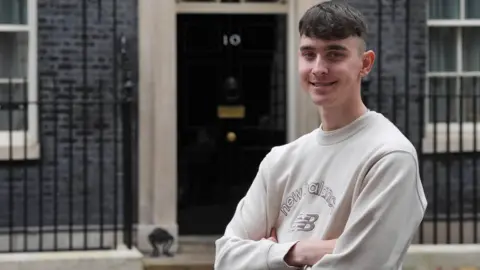 A young man smiling with his arms crossed standing in front of No 10 Downing Street