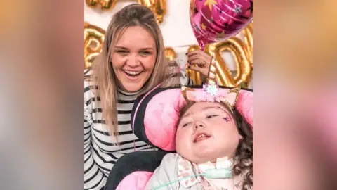 Family photograph Hayley Smallman with long blond hair wearing a white and black striped top smiles holding a metallic pink balloon and a gold happy birthday sign behind her with her young daughter Holly with long dark hair in curls wearing a unicorn headband and a pale pink dress. They are both smiling.