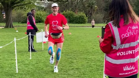 A man in a red British Heart Foundation shirt and shorts runs towards a parkrun volunteer dressed in a pink high visibility jacket. 