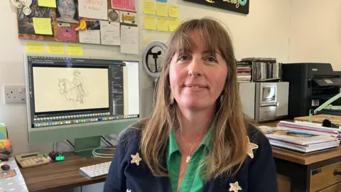 Rebecca Farkas sits in front of her work computer, with a hand-drawn image of a circus horserider visible on the screen. She is smiling and wearing a green shirt, and dark blue jumper with stars on. 