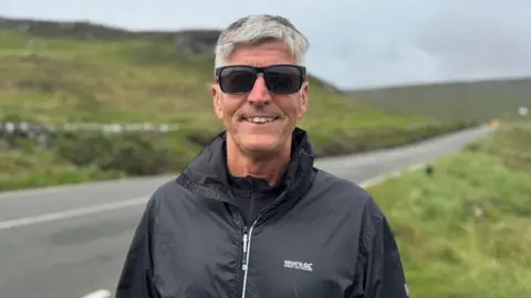 Andrew Titley has short grey hair, is wearing black sunglasses and wearing a black waterproof jacket, stood in front of a road lines with grass.