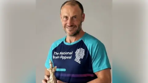 George Wade & Son Ltd A man with barely any hair and wearing a blue National Brain Appeal t-shirt smiles as he holds one of the small Ivy sculptures.