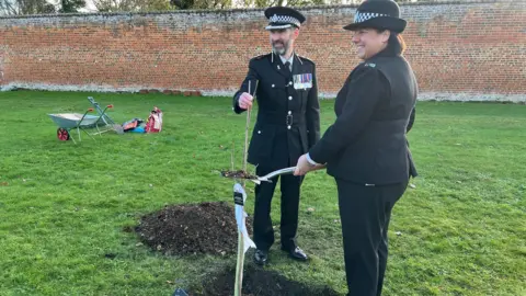 BBC Two police officers wearing formal uniforms stand over a hole in the ground where the sapling is being planted. One of the officers stands with a shovel over the hole. 
