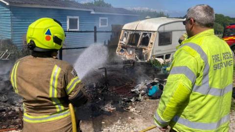 Two firefighters with one of them directing water at a smoking caravan