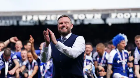 PA Media Mark Ashton is wearing a white shirt, navy blue knitted waistcoat and a striped tie. He is smiling and applauding, with Ipswich Town footballers celebrating behind him