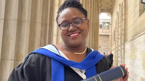 Police Scotland Dr Gomo, who is wearing black graduation robes, smiles as she holds a University of Dundee scroll. She is also wearing glasses.