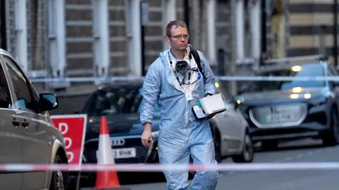 A man dressed in a blue forensic suit walks down the street. He has short brown hair and glasses, and there is a mask and lanyard around his neck. He looks somber. Behind him are parked cars and police tape.