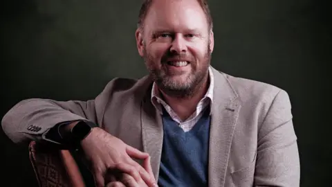 Ben Yearsley sitting on a chair, leaning an arm on the top if it. He wears a shirt, jumper and blazer and is smiling.