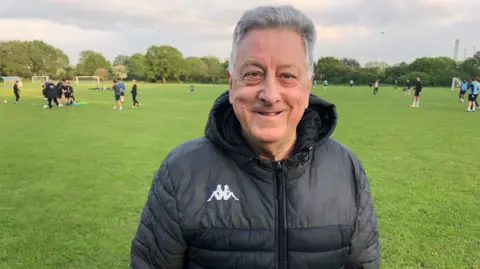 Jonathan Park/BBC David Patient, who is wearing a black coat and standing in front of people playing football