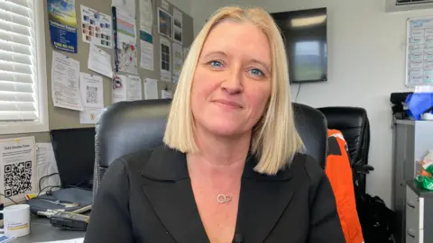 Justine Alexander has blonde hair and is wearing a black jacket. She is smiling for the picture as she sits in the current crew operations room. 
