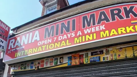 Rania Mini Market has a large sign with its name on in white writing, with a red background. Below the sign, the metal shop shutters are down.