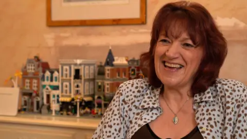 Natalie Zolty sits in front of a row of colourful houses constructed out of lego. She is smiling and has reddish hair cut into a bob, she is wearing a blue shirt with brown spots over a brown vest top. She is wearing a silver necklace with a blue and silver leaf-shaped pendant. 