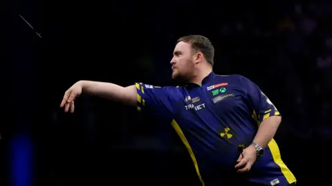 PA Media A side view of Luke Littler throwing a dart, wearing a blue and yellow darts shirt