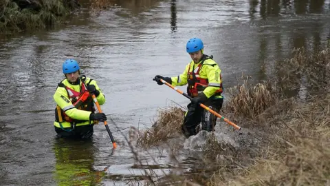 PA Two men wearing waterproof gear and blue helmets are standing in the river putting poles into it.