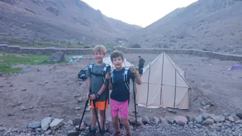 James Storey A bleak mountain landscape with Freddie and Max holding walking poles and wearing rucksacks, with two makeshift tents in the background