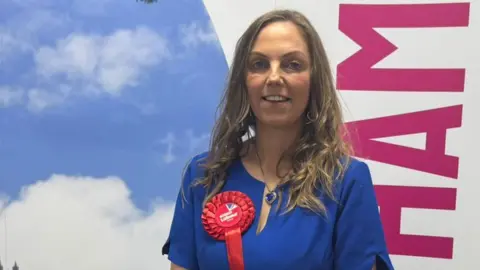 Julie Griffiths is smiling at the camera. She has long blonde wavy hair and blue eyes. She is wearing a short-sleeved blue dress with a red Labour rosette and blue heart pendant necklace. 