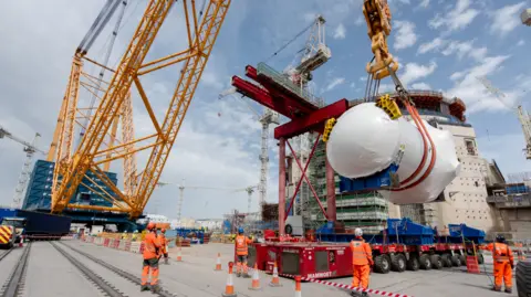 Hinkley Point C A giant yellow crane is lifting a large cylinder in white protective material.